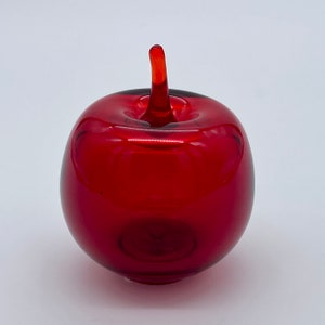 May include: A vibrant red glass apple sculpture with a stem. The apple is a solid, rounded shape with a glossy finish. The stem is a lighter shade of red, adding contrast to the design. This decorative piece is ideal for home decor.