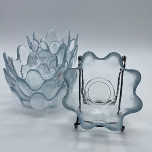Puede incluir: Un conjunto de cuencos de vidrio azul claro con un diseño único de borde festoneado. Un cuenco se muestra sobre un soporte de metal negro. Los cuencos tienen un aspecto esmerilado y están dispuestos sobre una superficie blanca.