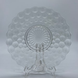 Puede incluir: Plato de vidrio transparente con un patrón de burbujas y borde festoneado. El plato tiene un centro circular con un diseño de estrella. Se muestra sobre un soporte de plástico transparente. El plato es probablemente vintage o antiguo.