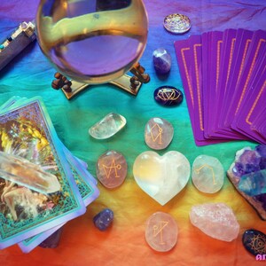 May include: A crystal ball, tarot cards, and various crystals are arranged on a rainbow-colored fabric. The crystals include a clear quartz point, a heart-shaped clear quartz, and several rose quartz stones with gold symbols. The tarot cards are purple with gold borders. The crystal ball is on a gold stand.