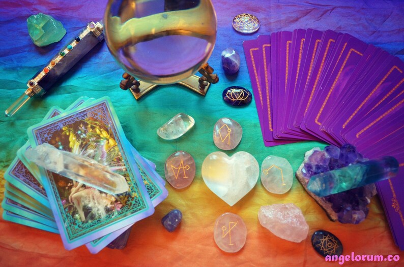 May include: A crystal ball, tarot cards, and various crystals are arranged on a rainbow-colored fabric. The crystals include a clear quartz point, a heart-shaped clear quartz, and several rose quartz stones with gold symbols. The tarot cards are purple with gold borders. The crystal ball is on a gold stand.