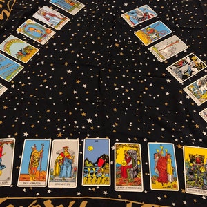 May include: A tarot card spread on a black fabric background with gold and white stars. The cards are arranged in a circle with the cards "The Hanged Man", "Page of Wands", "King of Cups", "Queen of Pentacles", "King of Cups", and "King of Pentacles" visible.