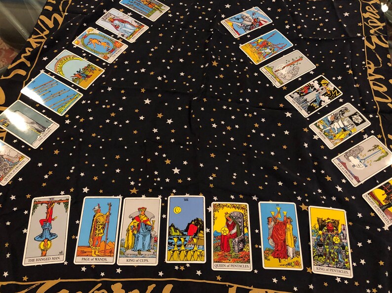 May include: A tarot card spread on a black fabric background with gold and white stars. The cards are arranged in a circle with the cards "The Hanged Man", "Page of Wands", "King of Cups", "Queen of Pentacles", "King of Cups", and "King of Pentacles" visible.