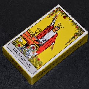 May include: A yellow and white tarot card with a red-robed figure holding a wand. The card is labeled "The Magician" and "Rider Waite Tarot".