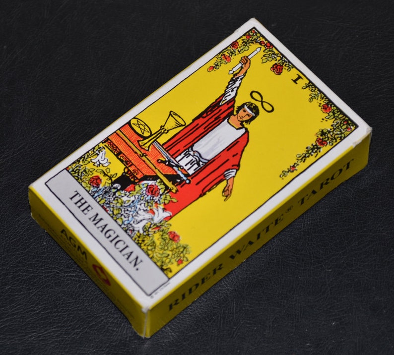 May include: A yellow and white tarot card with a red-robed figure holding a wand. The card is labeled "The Magician" and "Rider Waite Tarot".