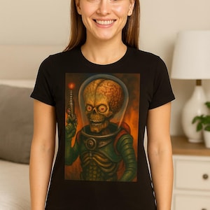 May include: Black crew neck t-shirt featuring a colorful illustration of a Martian holding a ray gun. The alien has a large brain, skull-like face, and is wearing a green space suit. The background is orange and red.