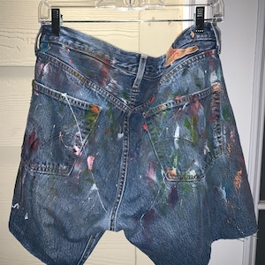 Puede incluir: Pantalones cortos de mezclilla azul con salpicaduras de pintura en varios colores, incluyendo rosa, verde, amarillo y naranja. Los pantalones cortos tienen un dobladillo deshilachado y están colgados en una percha.