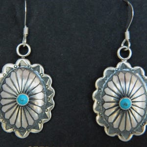 Op de afbeelding: Een paar zilverkleurige oorbellen met een turquoise middensteen. De oorbellen hebben een geschulpte rand en een radiaal ontwerp. De oorbellen zijn opgehangen aan een zilveren haak.