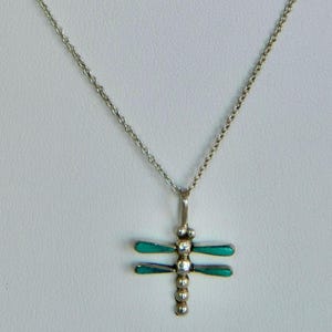 May include: A silver-toned necklace featuring a dragonfly pendant. The pendant body consists of a vertical row of small silver spheres, with two pairs of horizontal wings inlaid with smooth, vibrant teal-colored stones on each side.