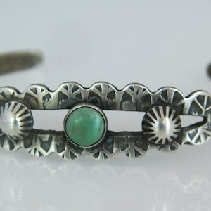 Puede incluir: Una pulsera de plata con una piedra central de color turquesa y dos diseños plateados en forma de sol. La pulsera tiene un borde festoneado decorativo y mide aproximadamente 6,35 cm de diámetro.