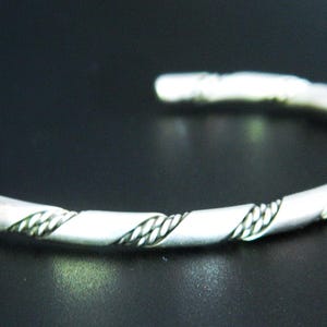 May include: A silver-colored cuff bracelet with a twisted rope design. The open-ended bracelet has a continuous pattern of interwoven lines, creating a textured appearance. The jewelry is displayed against a dark background.