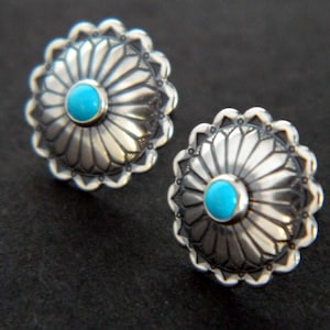 May include: A pair of silver earrings with turquoise stones set in a circular design. The earrings have a detailed, textured surface and a scalloped edge.