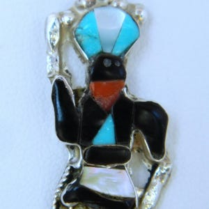 May include: A silver pendant necklace featuring a Native American Kachina doll made of turquoise, coral, and mother of pearl. The doll is wearing a blue headdress and has a black face.