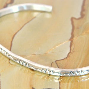 May include: A silver cuff bracelet with a repeating zigzag pattern along the top edge. The open-ended bracelet is displayed on a light brown and tan surface.