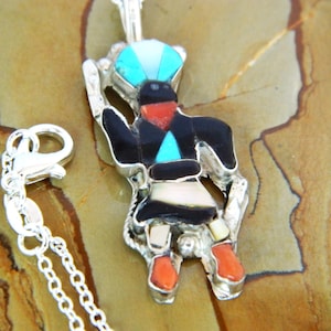 May include: A silver necklace with a Kachina doll pendant. The pendant is made of sterling silver and features turquoise, coral, and jet inlay. The doll is wearing a headdress and a skirt. The necklace is 18 inches long.