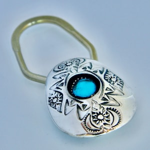 May include: A silver-toned metal keychain featuring a rounded, polished pendant with intricate stamped sun and crescent moon motifs. A vibrant, circular turquoise-colored stone is set in the center of the pendant, attached to a gold-toned loop.