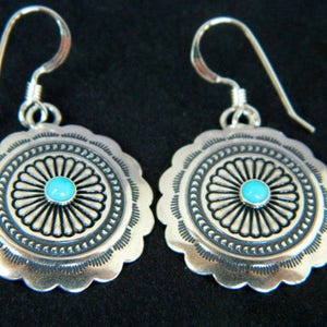 May include: A pair of silver earrings with turquoise stones set in a circular design. The earrings have a scalloped edge and a delicate wire hook.