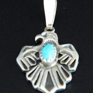 May include: A silver pendant shaped like an eagle with outstretched wings, featuring a central oval-shaped turquoise stone set in a serrated bezel. The metalwork includes cut-out details on the wings and tail, with a polished silver bail at the top.