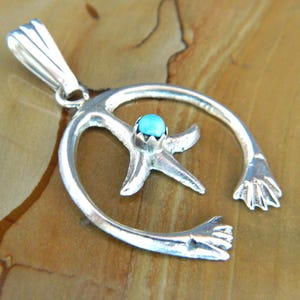 Puede incluir: Colgante de plata con una estrella de mar con una gema turquesa en el centro, rodeada por un diseño de ola estilizada. El colgante tiene una anilla para una cadena y dos formas de mano en los extremos.