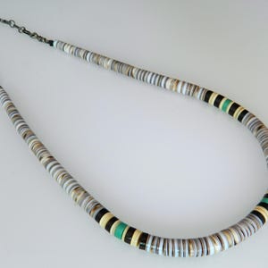 Puede incluir: Un collar compuesto por cuentas planas y redondas en tonos blanco, gris, marrón y negro. Las cuentas están ensartadas, con una sección de cuentas verdes. El collar está conectado por una cadena de color plateado.