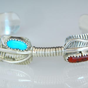 May include: Silver cuff bracelet with feather design. Features a turquoise stone and a red stone. The bracelet has an open design and is likely made of sterling silver.