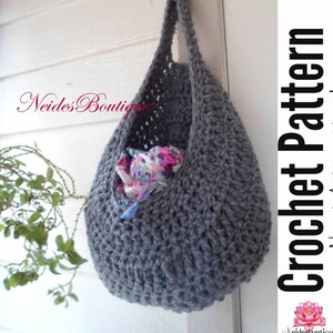 Op de afbeelding: Een grijze gehaakte hangmand met een handvat, gevuld met een kleurrijke bloemenstof. De mand hangt aan een wit oppervlak. De tekst "Crochet Pattern" staat aan de rechterkant van de afbeelding.