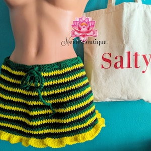 Pu&ograve; includere: Una gonna a crochet a righe verdi, nere e gialle con un bordo di volant gialli. La gonna &egrave; su un manichino e ha una chiusura con coulisse. Una borsa tote bianca con la parola "Salty" stampata in rosso &egrave; sullo sfondo.