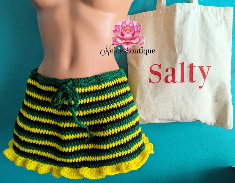Pu&ograve; includere: Una gonna a crochet a righe verdi, nere e gialle con un bordo di volant gialli. La gonna &egrave; su un manichino e ha una chiusura con coulisse. Una borsa tote bianca con la parola "Salty" stampata in rosso &egrave; sullo sfondo.