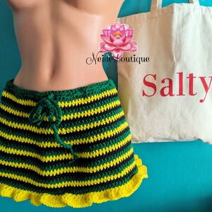 Pu&ograve; includere: Una gonna a crochet a righe verdi, nere e gialle con un bordo di volant gialli. La gonna &egrave; su un manichino e ha una chiusura con coulisse. Una borsa tote bianca con la parola "Salty" stampata in rosso &egrave; sullo sfondo.