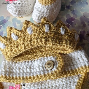 May include: A crocheted baby outfit in white and gold with a crown hat, booties, and a button closure. The outfit is made with a soft yarn and is perfect for a newborn baby.
