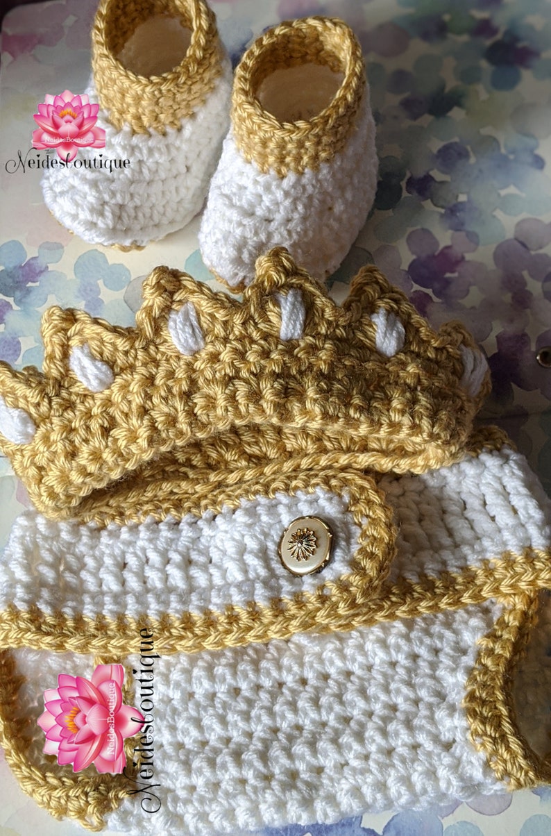 May include: A crocheted baby outfit in white and gold with a crown hat, booties, and a button closure. The outfit is made with a soft yarn and is perfect for a newborn baby.