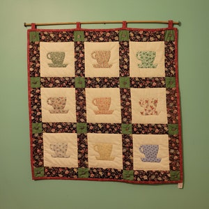 May include: A quilted wall hanging with a floral pattern featuring nine teacups in various colors and patterns.