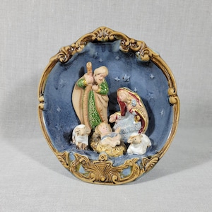 May include: A blue and gold ceramic wall plaque depicting the nativity scene. The plaque features a three-dimensional depiction of Mary, Joseph, and baby Jesus with two sheep.