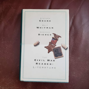 May include: A white book with a teal border titled "The Civil War Reader: Literature" with the names Stephen Crane, Walt Whitman, and Ambrose Bierce printed on the cover. The cover also features a vintage photograph of a soldier and a metal star with an American flag.