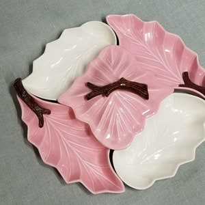 May include: A set of four ceramic leaf-shaped serving dishes with brown twig handles. The dishes are in a pink and white color scheme.