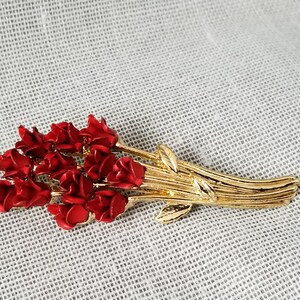 May include: A gold-toned brooch featuring a bouquet of red roses. The roses are clustered together and have a realistic appearance. The stems are gold-toned and have leaves.