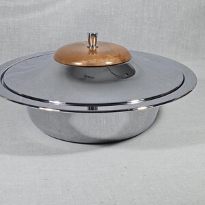 May include: A silver-colored serving bowl with a lid. The lid has a wooden, circular handle. The bowl is round with a wide rim and sits on a light-colored surface. The lid is also silver with a wooden top.