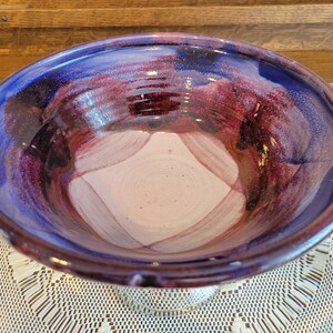 May include: A large, ceramic serving bowl with a deep, rounded shape. The bowl is glazed in shades of purple, blue, and pink, with a swirling, abstract design.