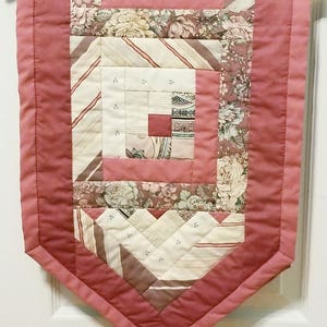 May include: A pink and white patchwork table runner with a geometric design. The runner is made of various fabrics with floral patterns and stripes. It has a pointed end and is hanging from a wooden hanger.