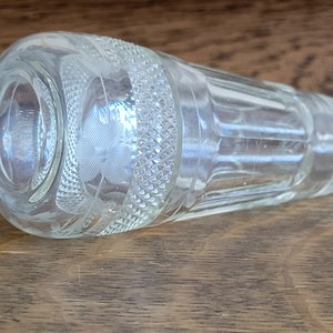 May include: A clear glass bottle with a textured surface and a decorative etched design. The bottle is lying on a wooden surface.