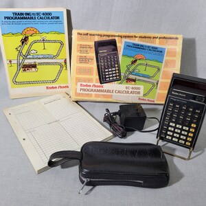 May include: A vintage Radio Shack EC-4000 programmable calculator with its original box, instruction manual, and carrying case. The calculator has a black and silver design with a small LCD screen. The box is yellow and blue with a cartoon illustration of a train on a track. The manual is titled "Training the EC-4000 Programmable Calculator".