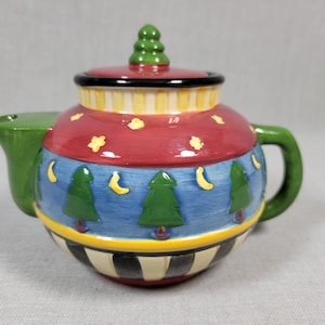 May include: A ceramic teapot with a green lid and handle. The teapot is decorated with a red, blue, yellow, and green pattern of trees, moons, and stars.