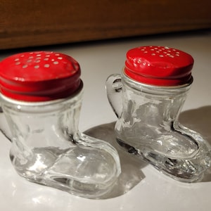 May include: Two clear glass salt and pepper shakers shaped like boots with red lids. The lids have a small hole in the center and a pattern of small white dots.