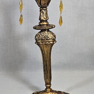 May include: A gold-toned table lamp with a decorative base and a finial. The lamp has three dangling crystal prisms.