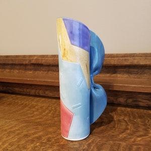 May include: A ceramic vase with a blue, purple, yellow, and red geometric design. The vase has a unique shape with a curved, blue section.