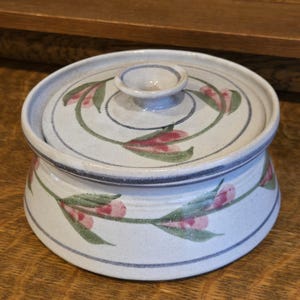 May include: A round, off-white ceramic casserole dish with a matching lid, featuring a hand-painted pattern of green vines and pink flower buds encircling both the base and the lid. The piece has a smooth, speckled finish and a central knob handle.