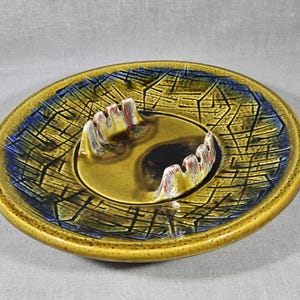 May include: A vintage ceramic ashtray with a circular design. The ashtray features a yellow-green base with a blue and black geometric pattern around the rim. The center has a dark brown area with raised rests for cigarettes.
