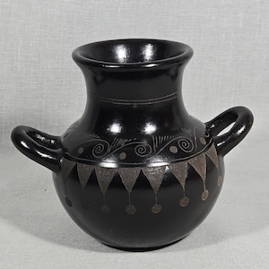 May include: A small, glossy black ceramic vessel with two side handles and a wide, flared rim. The body features etched decorative patterns, including a row of downward-pointing triangles with circular accents and swirling line motifs above.