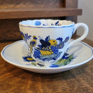 May include: A white teacup and saucer with a blue and yellow floral design. The design features birds, flowers, and fruit.