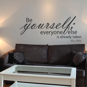 May include: A black and white wall decal with the quote "Be yourself; everyone else is already taken - Oscar Wilde". The decal is on a white wall above a brown sofa and a glass-topped coffee table.